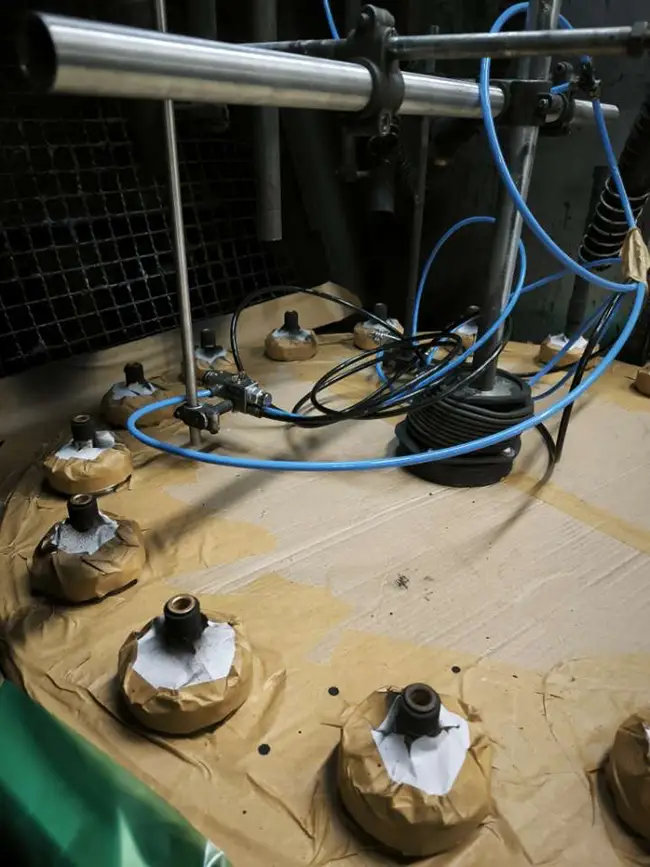 Vue rapprochée d'un plateau rotatif de cabine de peinture industrielle, avec plusieurs pièces mécaniques protégées par du papier kraft et fixées sur des supports, reliées par des tuyaux pneumatiques bleus.