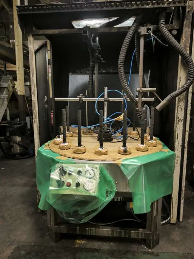 Vue d'ensemble d'une cabine de peinture industrielle avec un plateau tournant recouvert d'une bâche verte, sur lequel sont disposées des pièces à peindre montées sur tiges, avec le boîtier de commande protégé par un film plastique transparent.